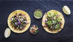 top down photo of two authentic mexican street tacos on slate surface with onions, pico de gallo, salsa verde, limes, cilantro, and jalapeno peppers